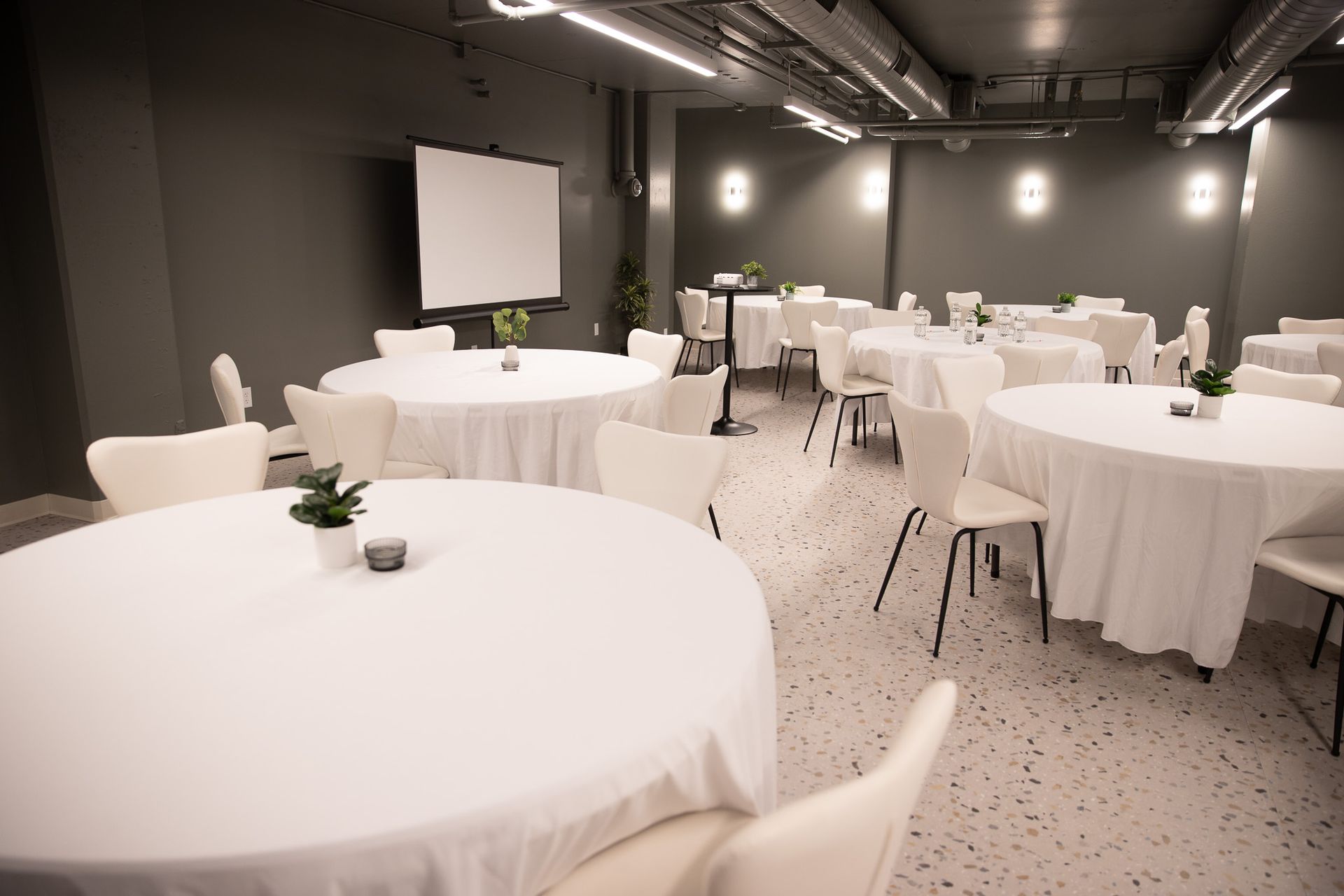 A modern, dim-lit event room with multiple round tables draped in white cloths, white chairs, and a projector screen.