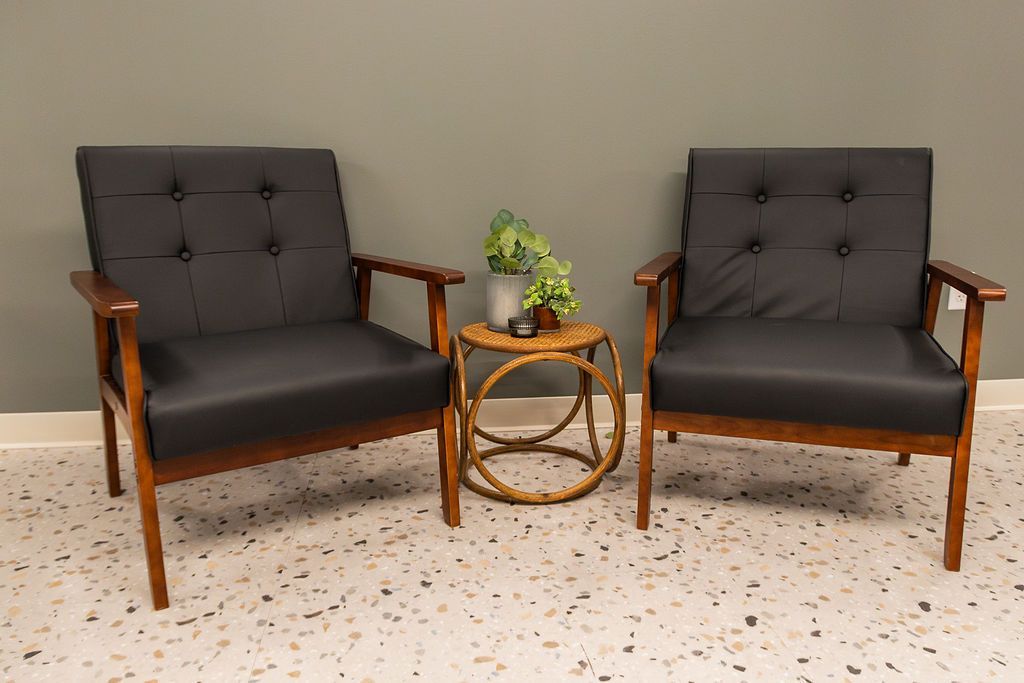 Two black tufted armchairs with dark wood frames sit on a speckled floor, separated by a small, round wicker table.
