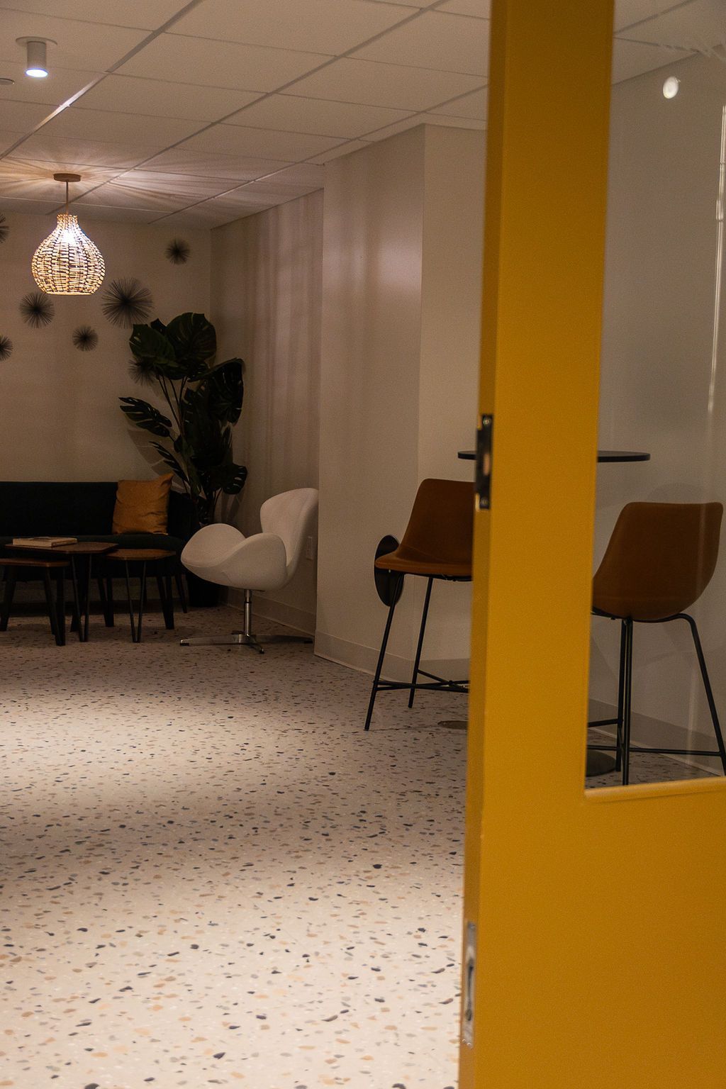 A view through a yellow door frame into a modern lounge with terrazzo floors, a white accent chair, and two bar stools.