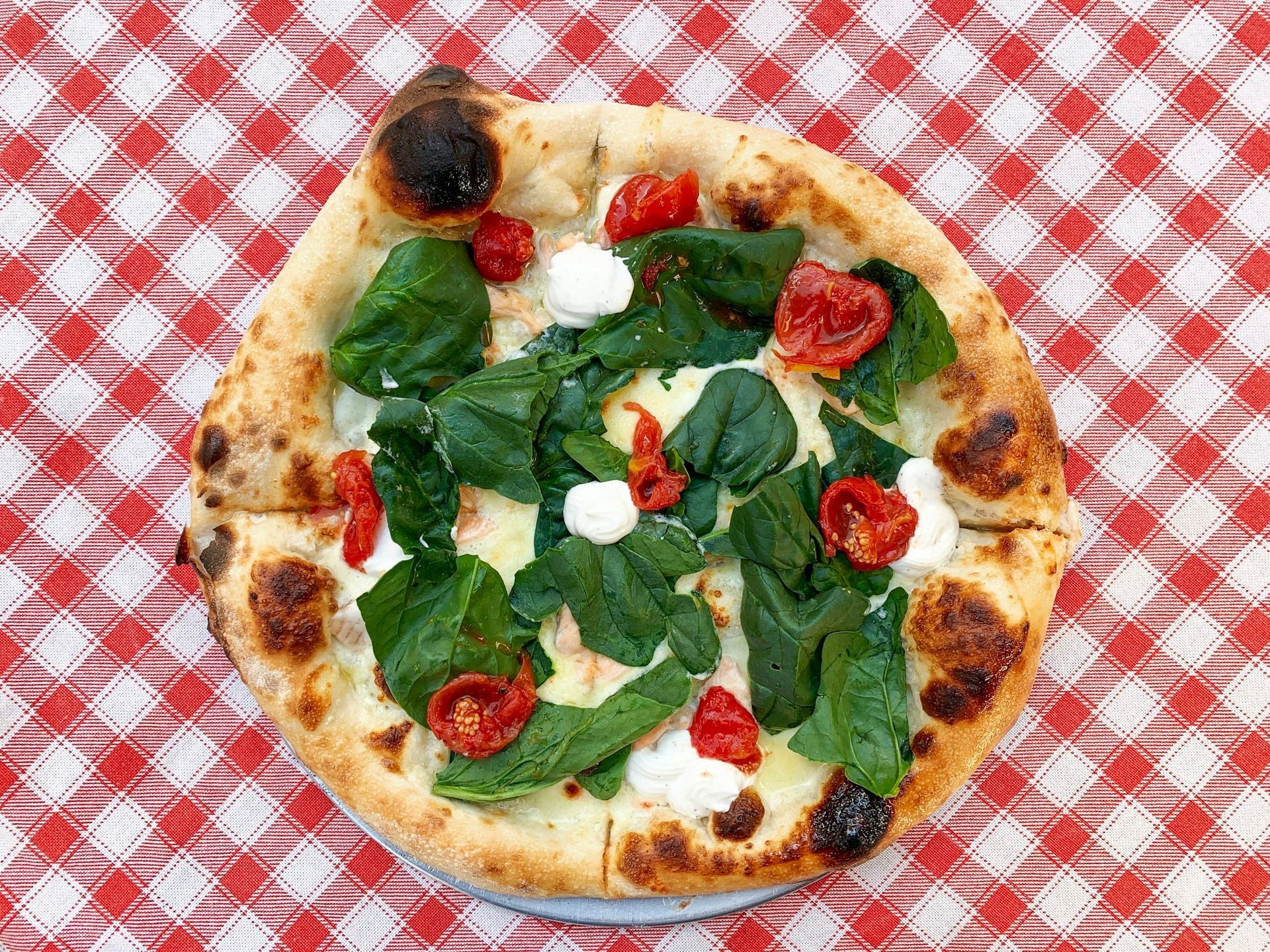 A gourmet pizza topped with fresh spinach, dollops of ricotta, and roasted cherry tomatoes on a red-checkered tablecloth.