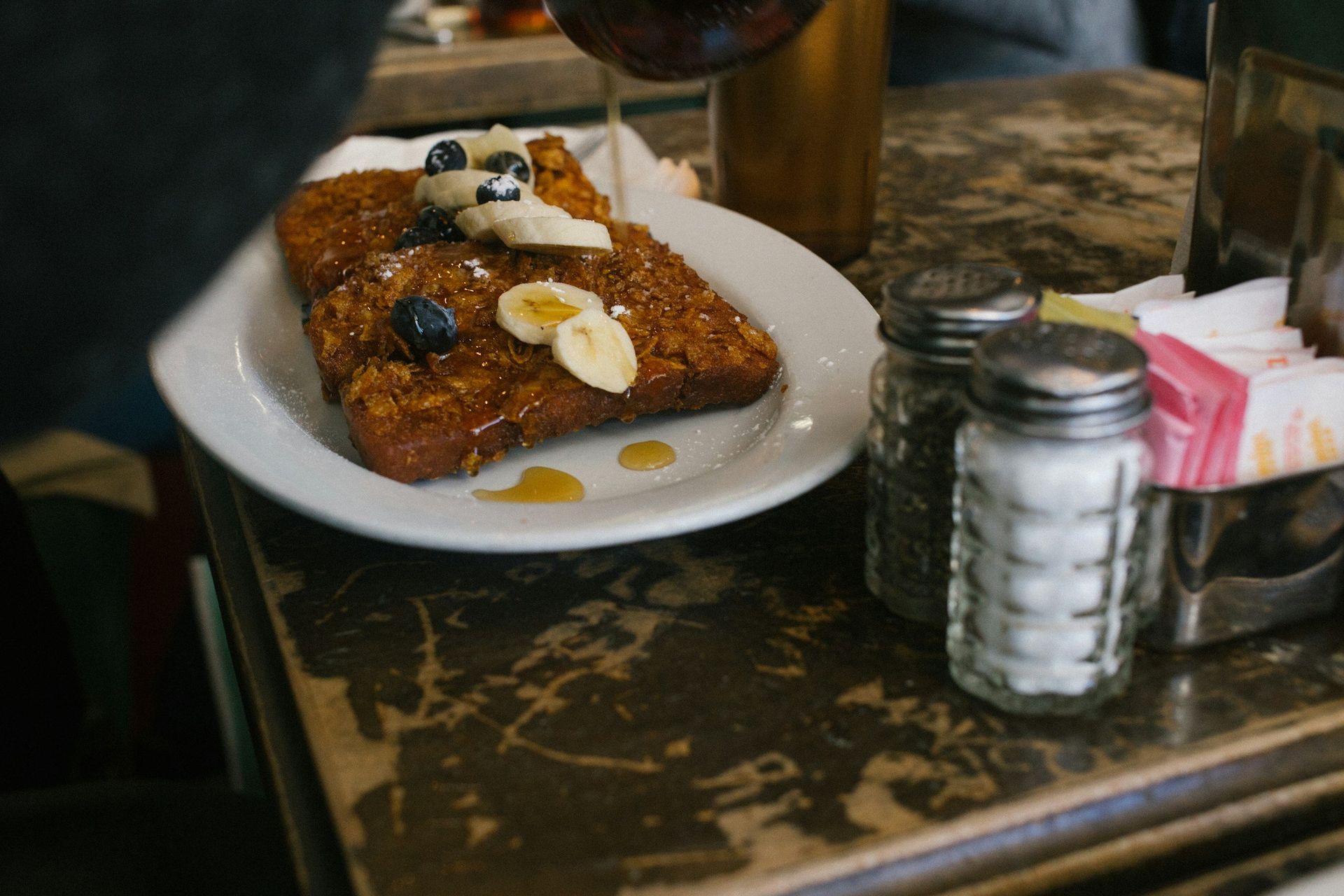 A slice of French toast topped with blueberries and banana slices on a white plate, with syrup being drizzled over it.