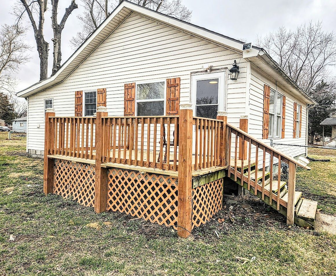 A small, one-story white house with light-colored siding, a wooden front deck, and lattice skirting under the porch.