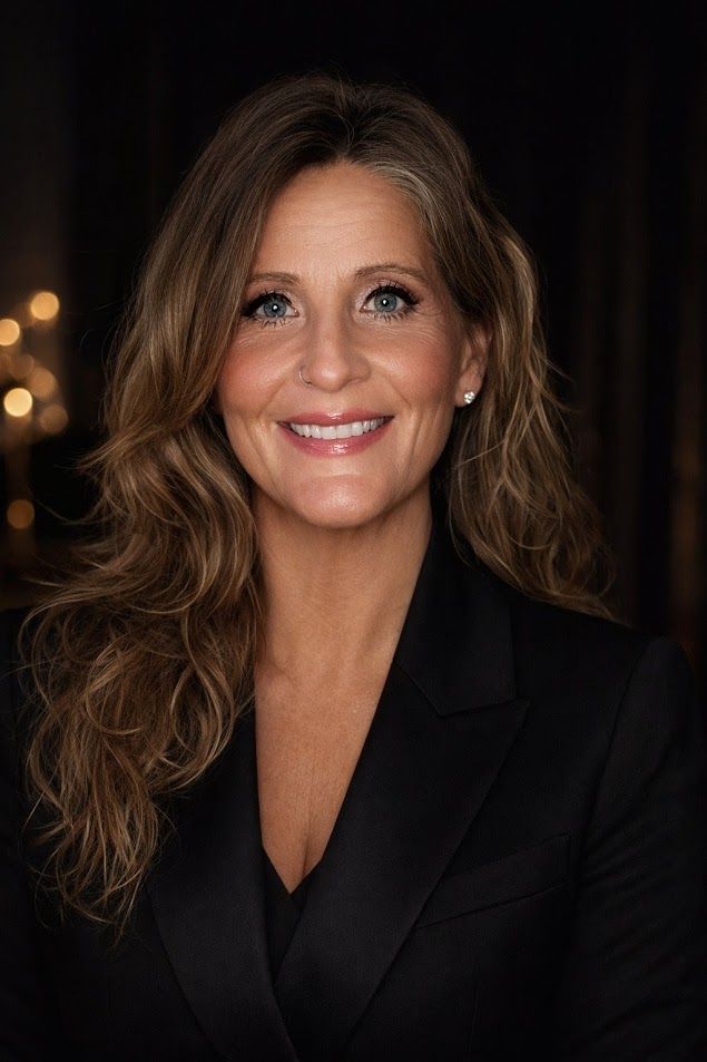 A person with long brown hair, wearing a black blazer, smiles while facing forward against a dark, softly lit background.