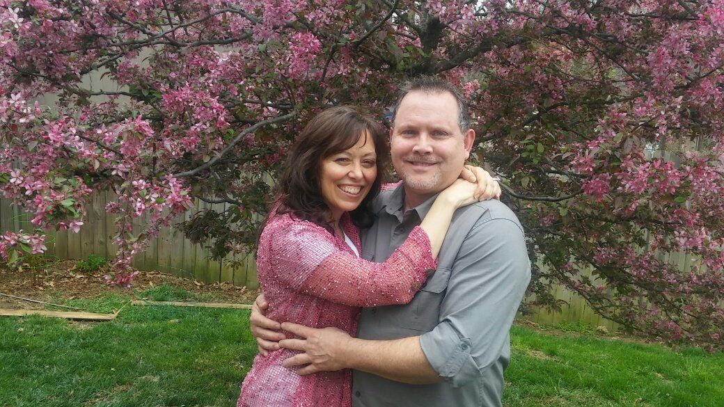 A man and a woman are hugging in front of a tree with pink flowers.