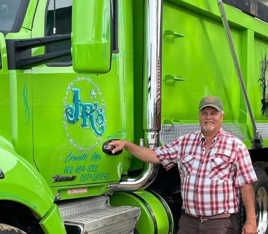 A man in a plaid shirt is standing next to a green dump truck.