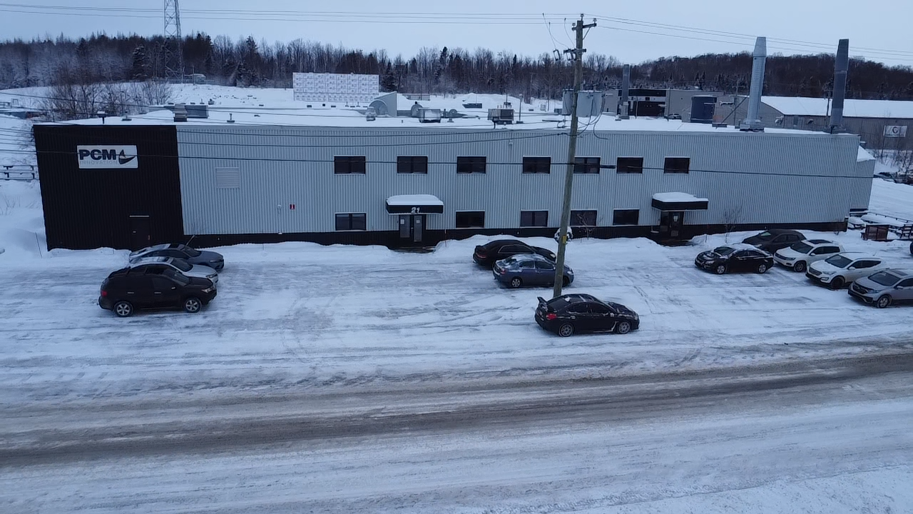 Vue extérieure d'un bâtiment commercial avec des voitures garées sur un parking enneigé. Bâtiment gris et noir en hiver.