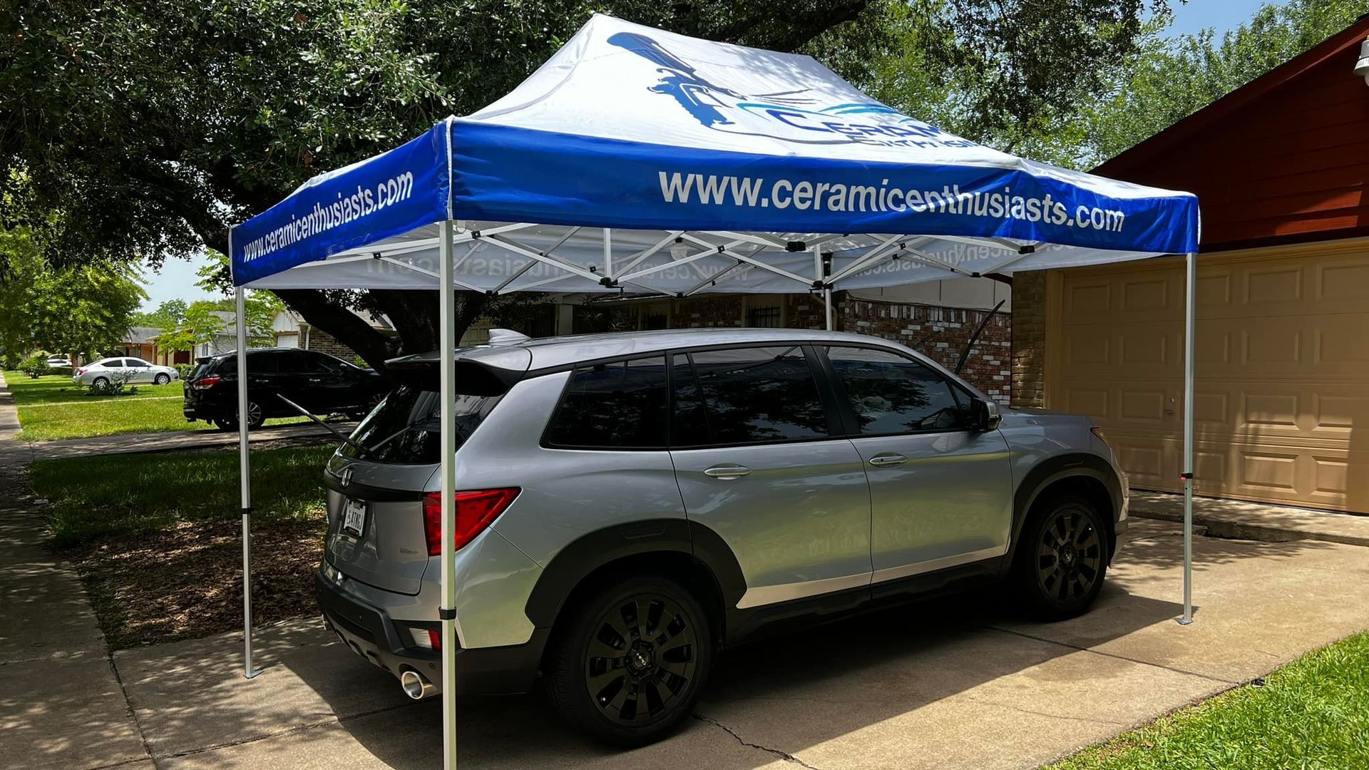 Silver SUV under a blue and white tent. Tent has company logo and website. Parked in front of a house.