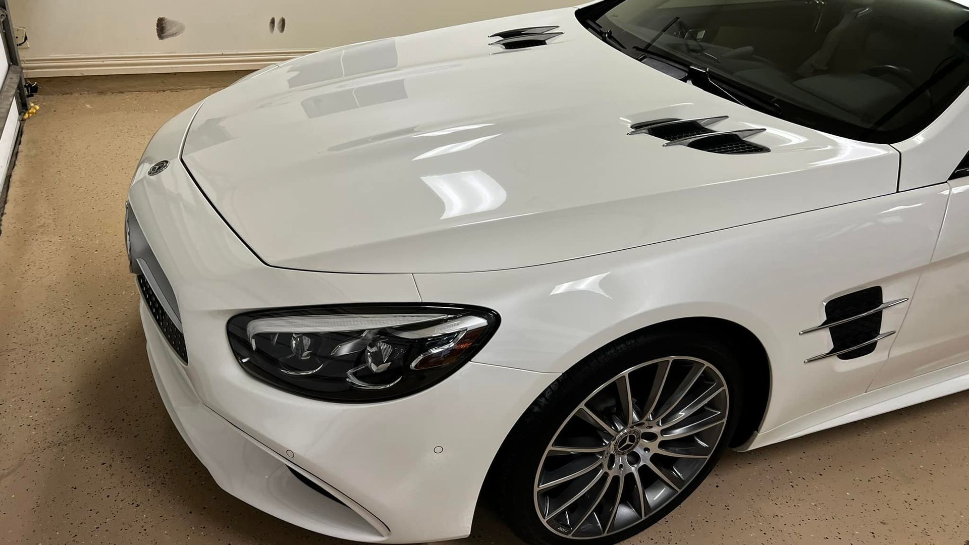 White Mercedes-Benz convertible parked indoors, displaying sleek design, visible front tire and hood vents.