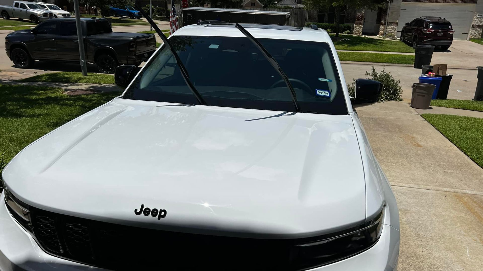 White Jeep parked on a driveway with windshield wipers up. Black Jeep and trash cans are in the background.