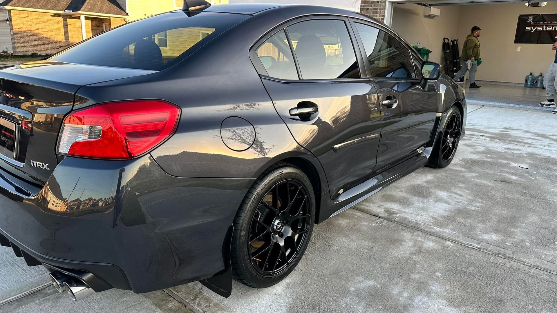 Dark gray Subaru WRX parked, black wheels, tinted windows. People in background near a garage.