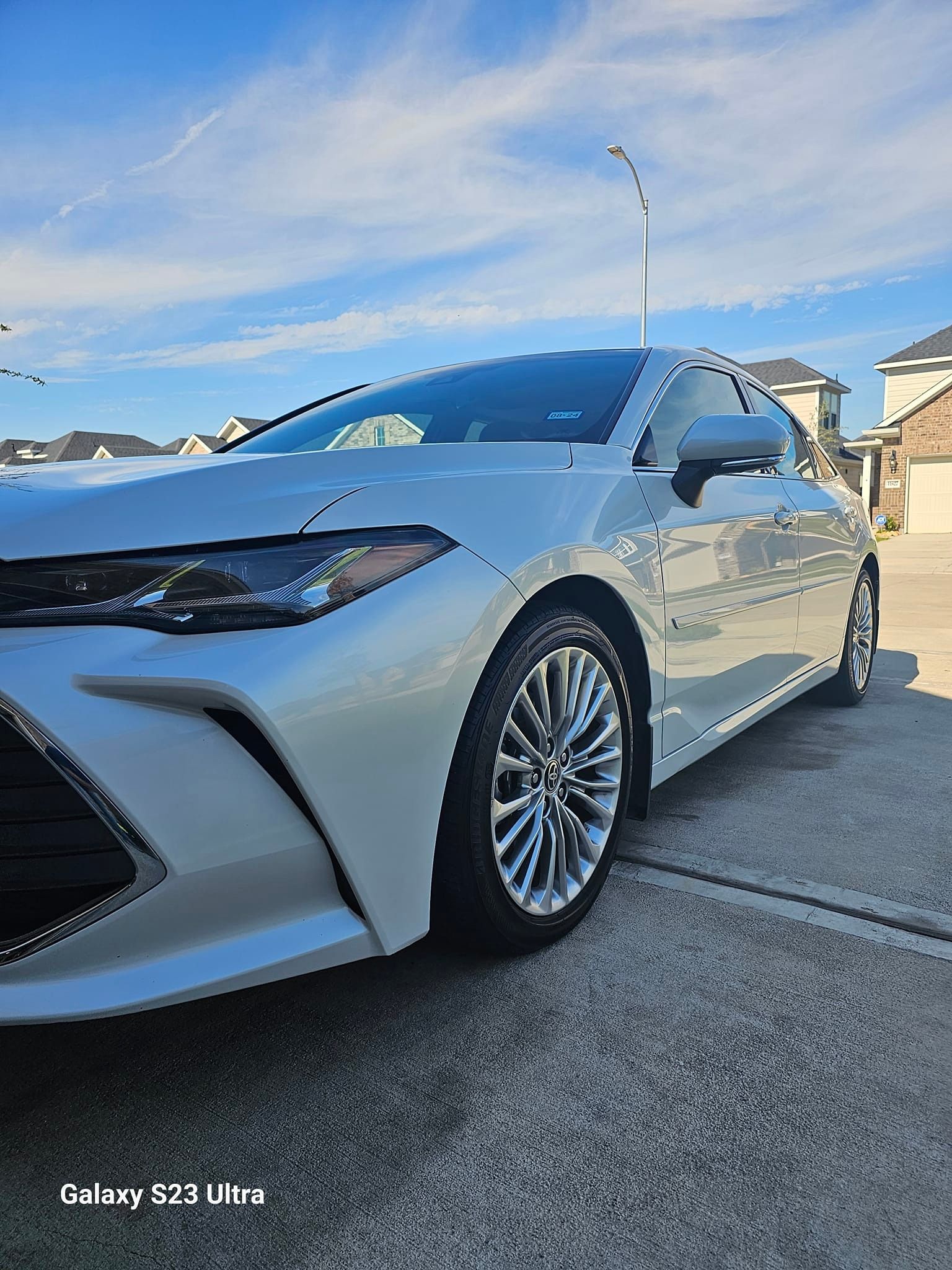 White Toyota Avalon parked on a concrete driveway under a blue sky with light clouds.