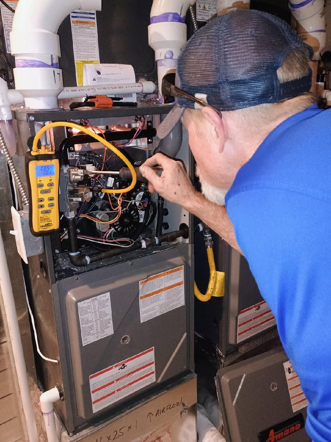 Person inspecting a furnace with a yellow multimeter. Inside a utility room with pipes and ventilation.