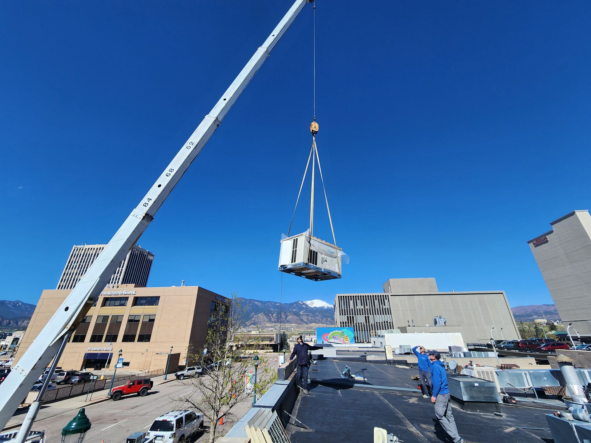 A crane lifting an HVAC unit onto a building rooftop on a sunny day.