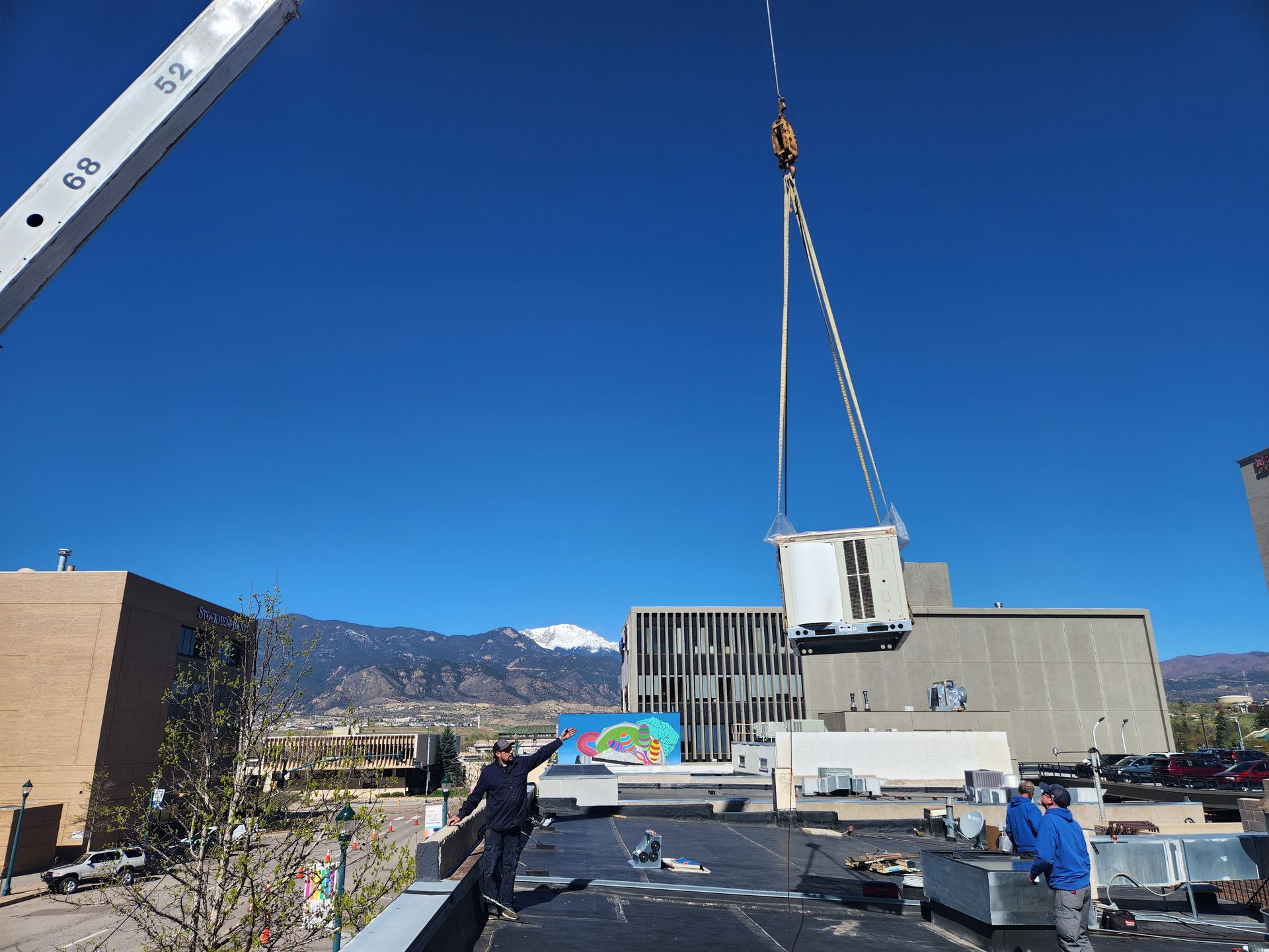 A crane lifts an HVAC unit onto a building roof under a blue sky, with workers present. Mountain in the background.