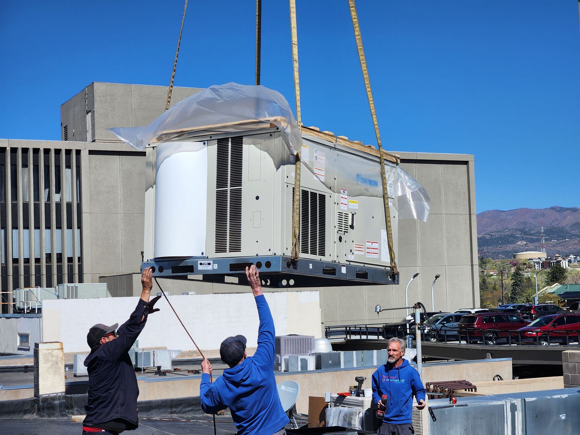 HVAC unit being lifted by crane onto a building rooftop; three workers direct the load, clear sky.