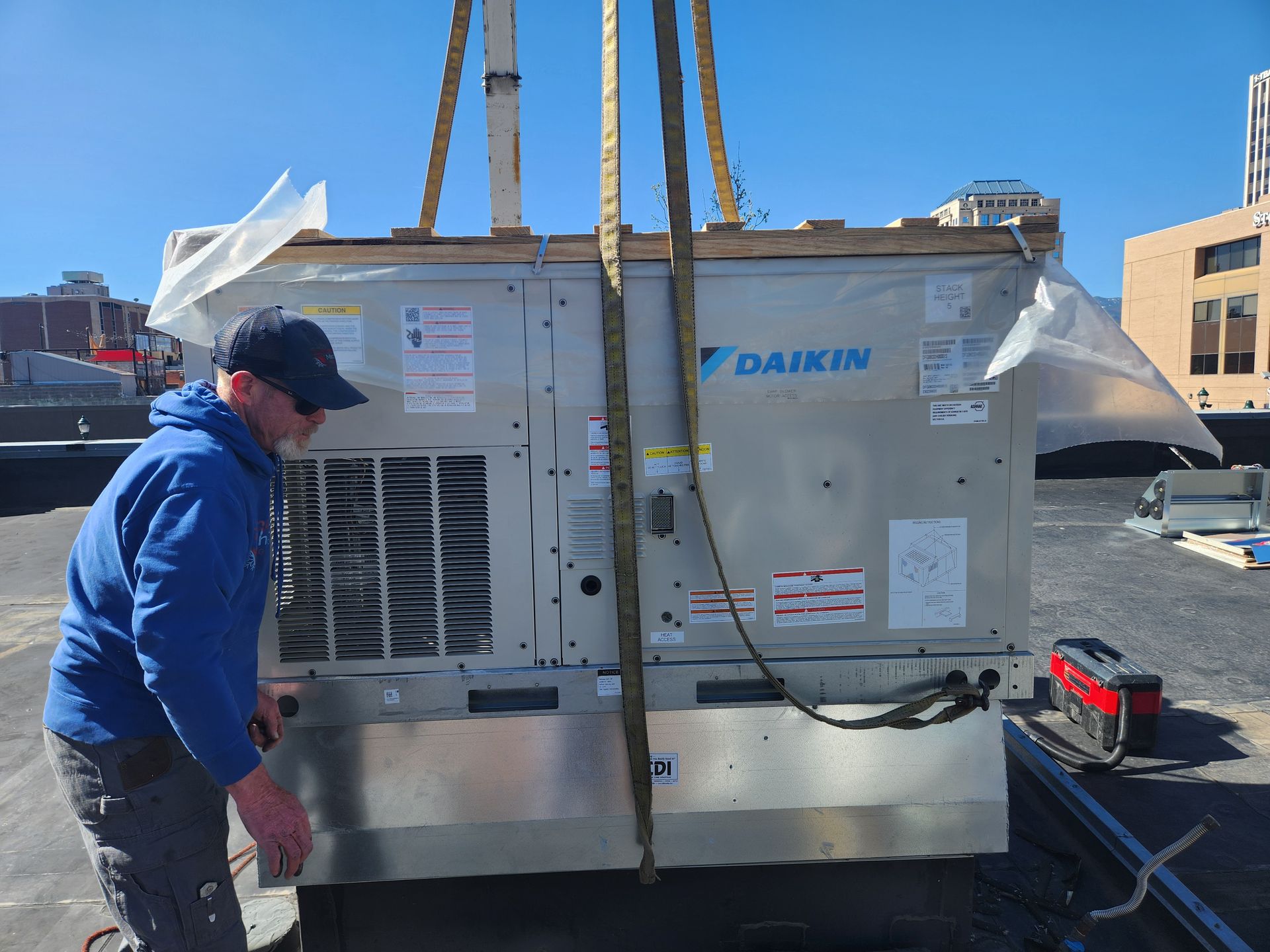 HVAC unit being lifted by straps onto a rooftop. A person in blue adjusts the unit. Clear sky.