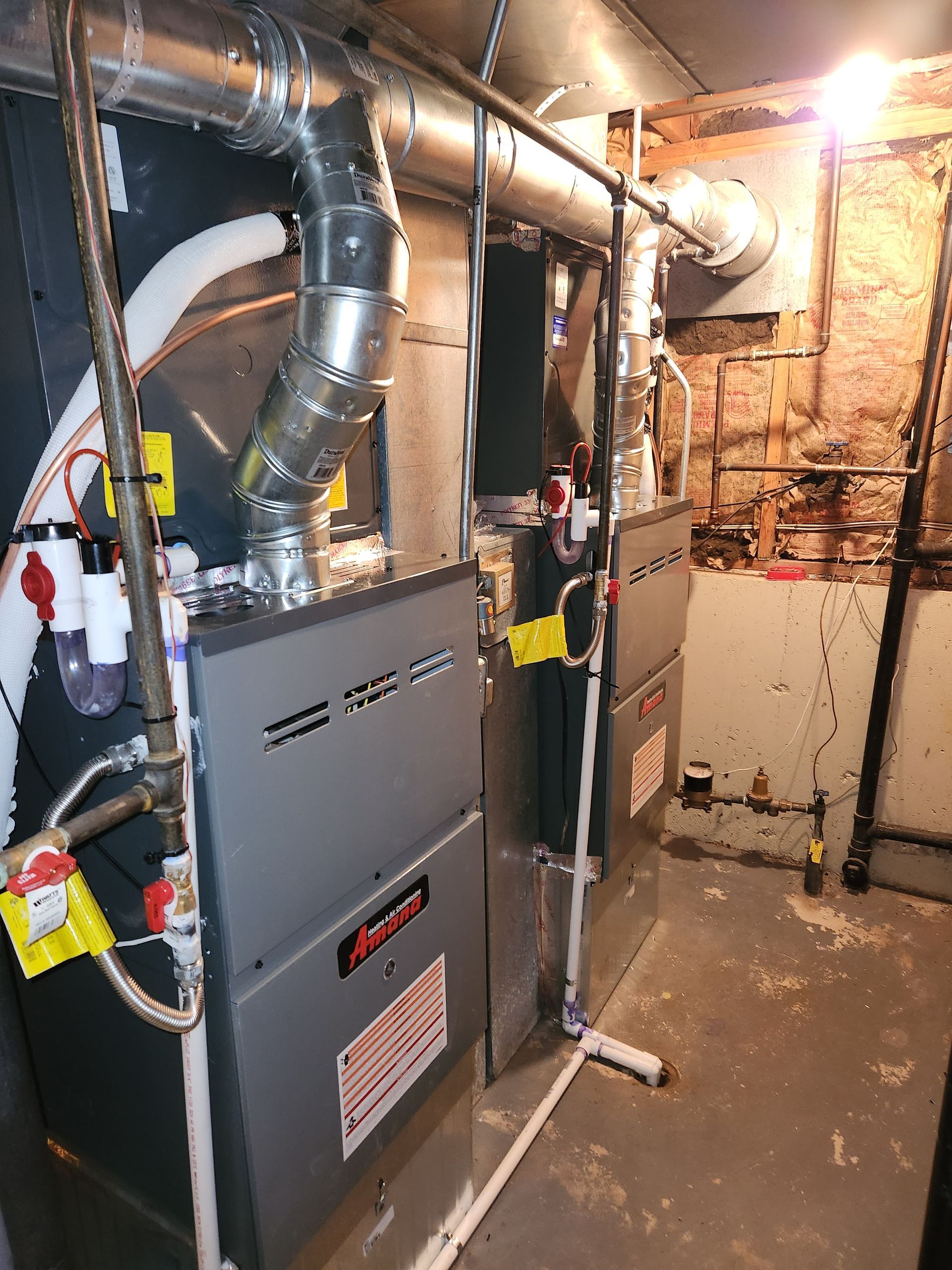 Two gray furnaces with silver ductwork in a basement with exposed brick and concrete floor.