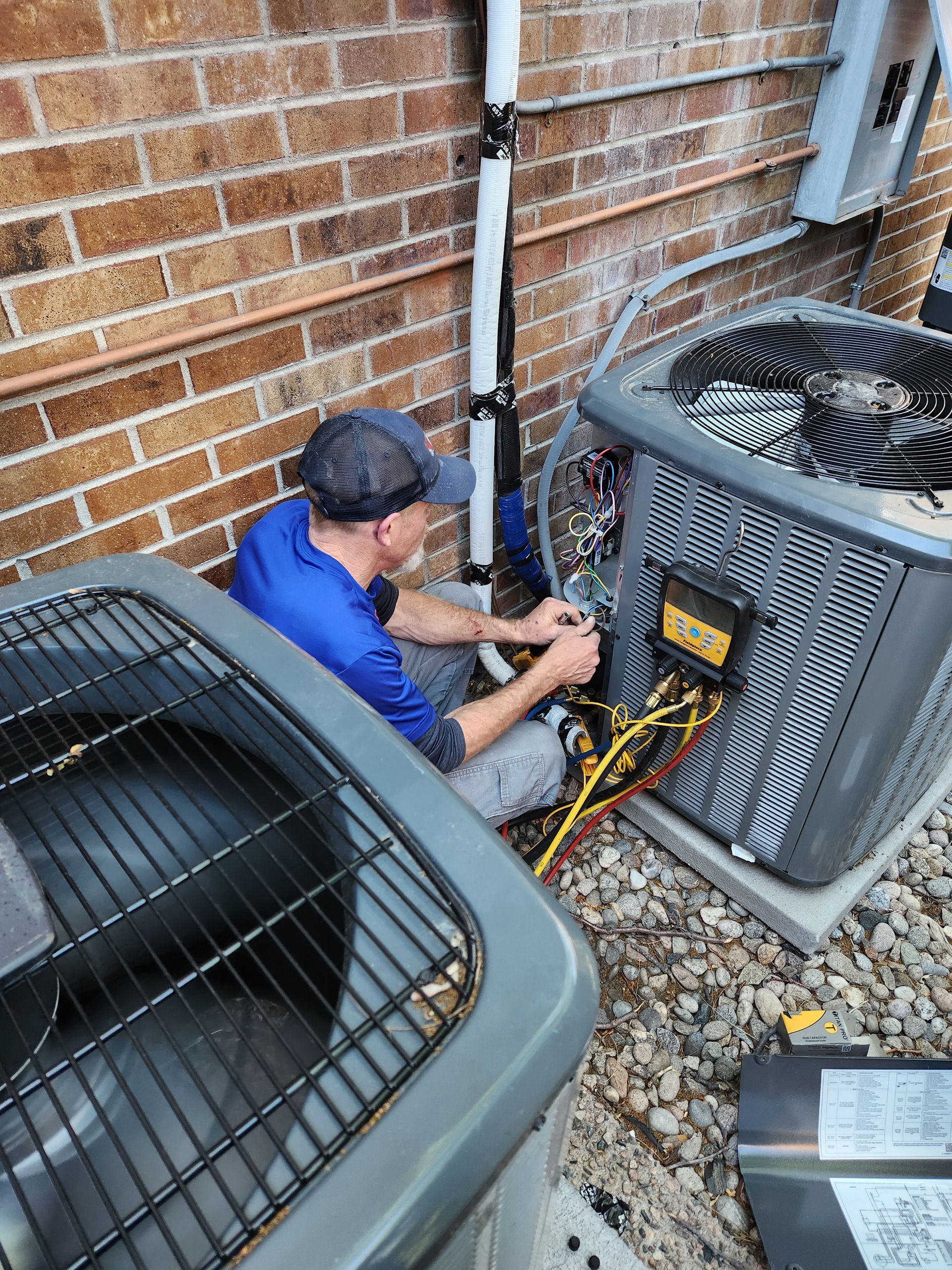 HVAC technician working on outdoor air conditioning units near a brick wall, wires visible.