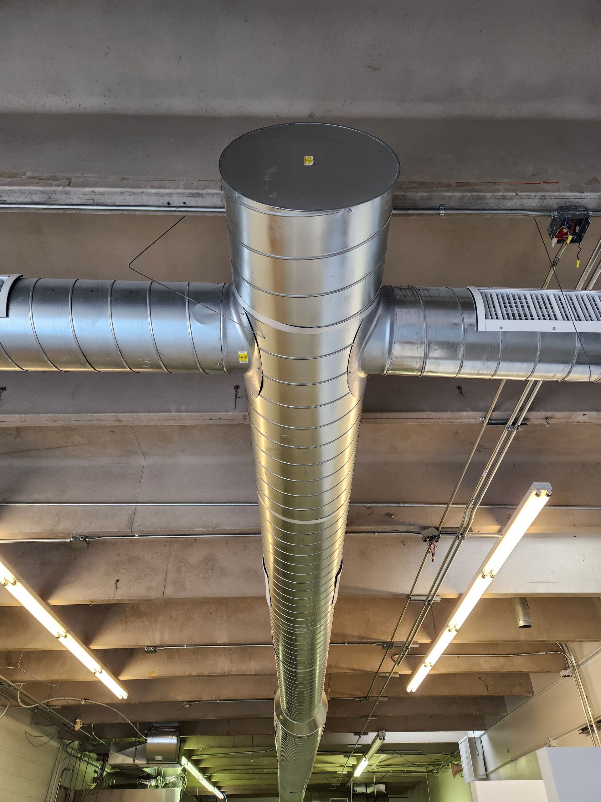 Metal HVAC ductwork system on a concrete ceiling, with horizontal and vertical tubes, and overhead fluorescent lights.