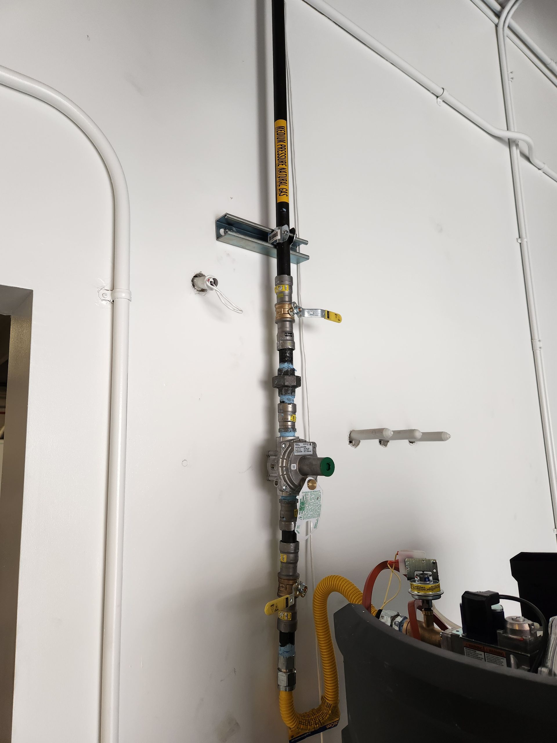 Vertical gas piping against a white wall with brackets, valves, and a yellow flexible connector.