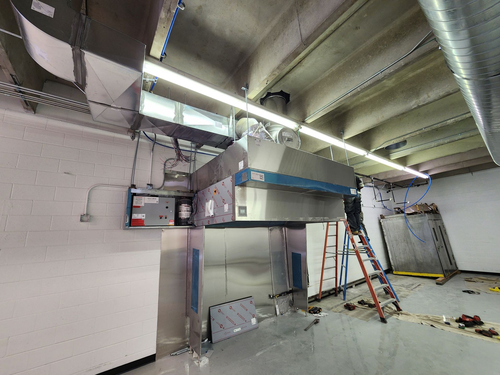 Commercial kitchen ventilation hood being installed. Shiny metal and gray ductwork against white walls and ceiling.