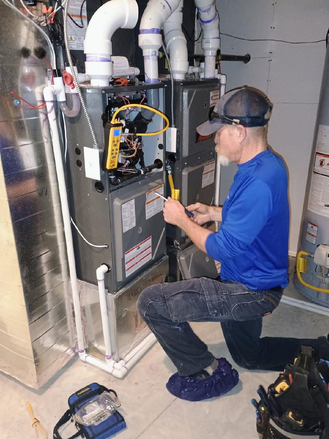 HVAC technician kneeling, inspecting a furnace. Wearing blue shirt, ball cap, and protective shoe covers. Interior setting.