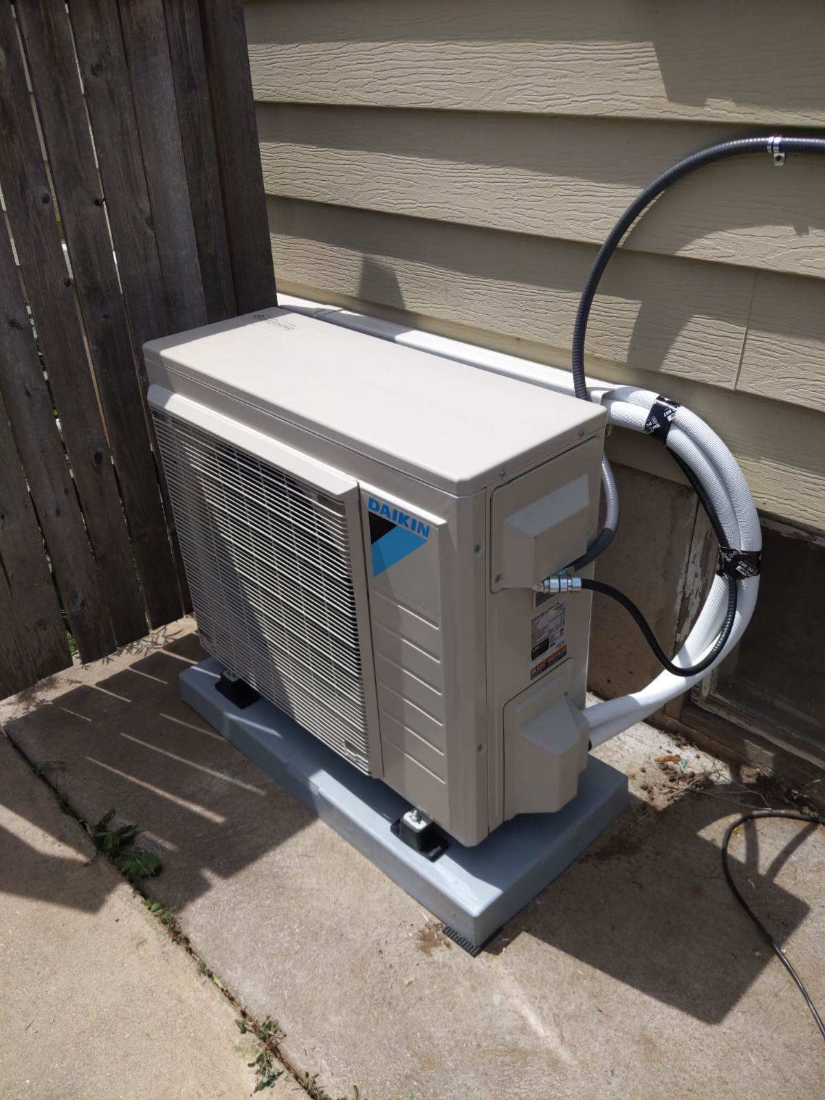 An outdoor air conditioning unit, light tan with black and white tubing, sits on a gray concrete