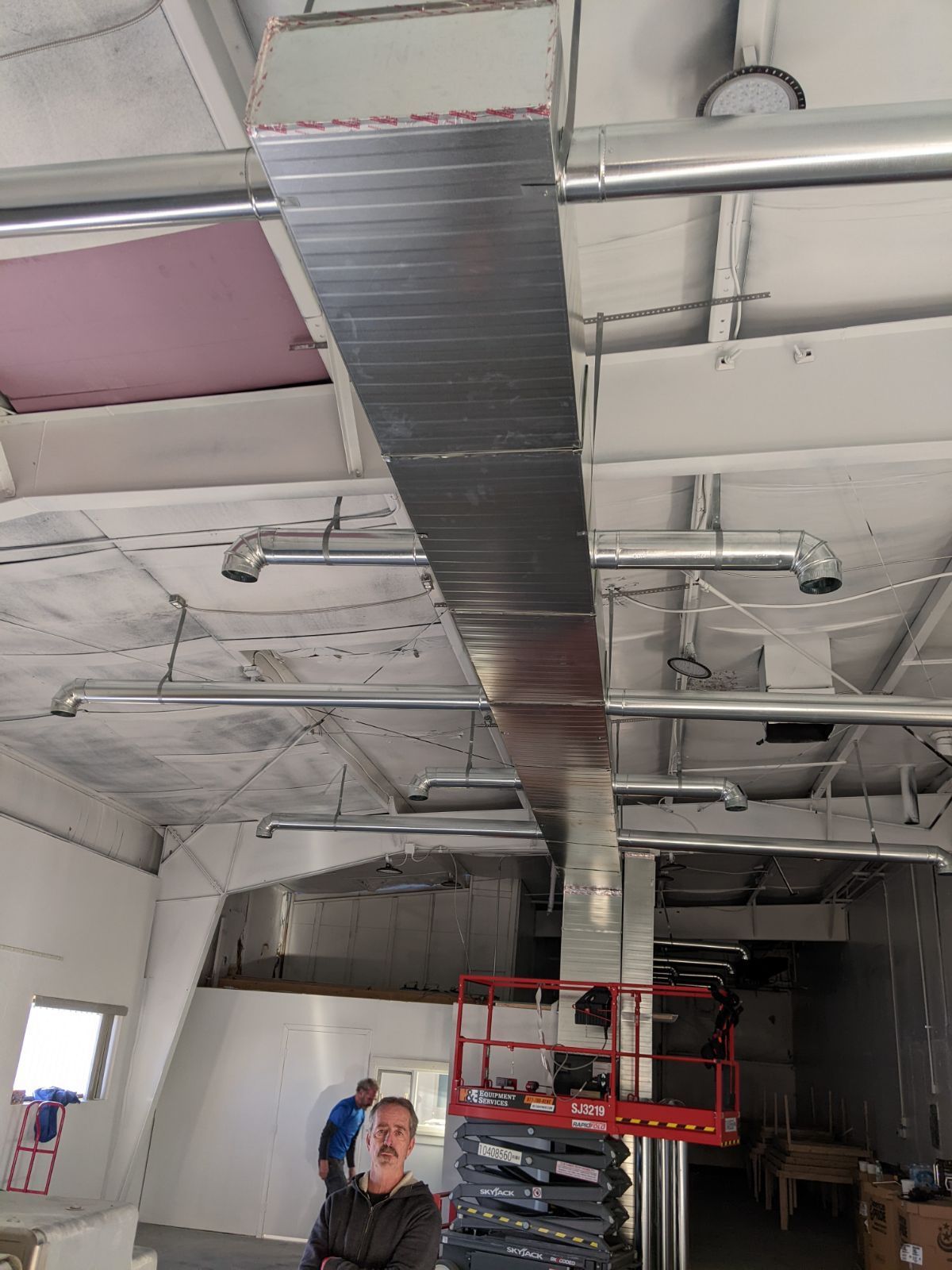 A long rectangular metal duct hangs from a white ceiling, with some workers and a scissor lift below.