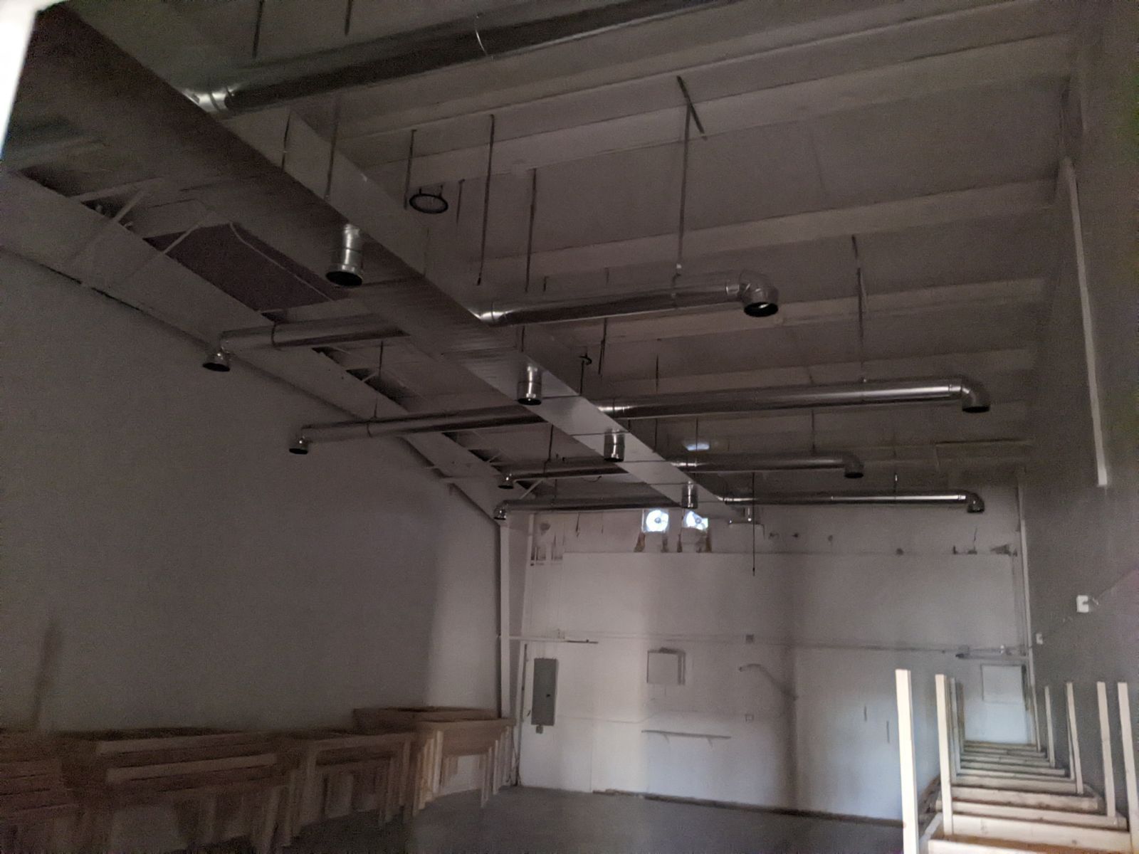 Interior of a room with exposed ductwork on the ceiling. White walls, a table, and a ladder are visible.