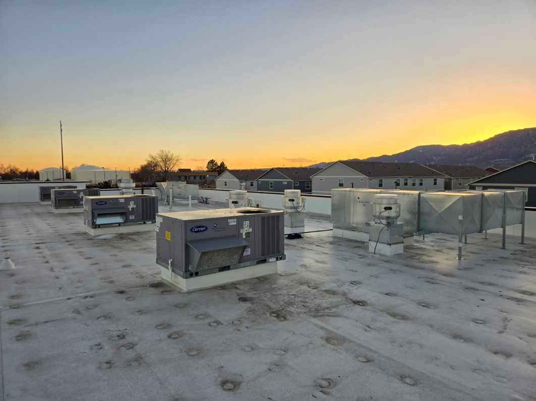 Rooftop with HVAC units against a sunset. Buildings and mountains are visible in the distance.