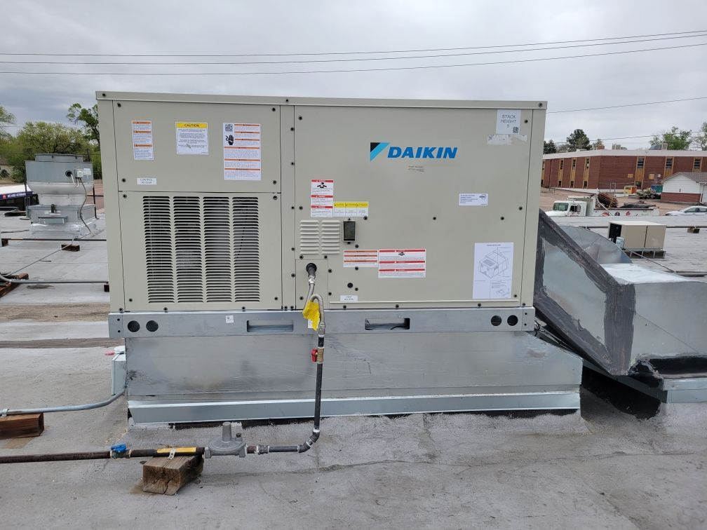 Daikin HVAC unit on a flat roof. Beige unit with a silver base, metal ductwork, and pipes. Cloudy sky background.
