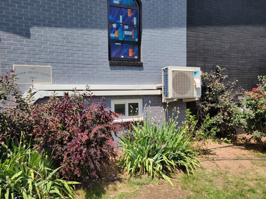 Exterior of a brick building with a stained glass window, surrounded by bushes and an air conditioning unit.
