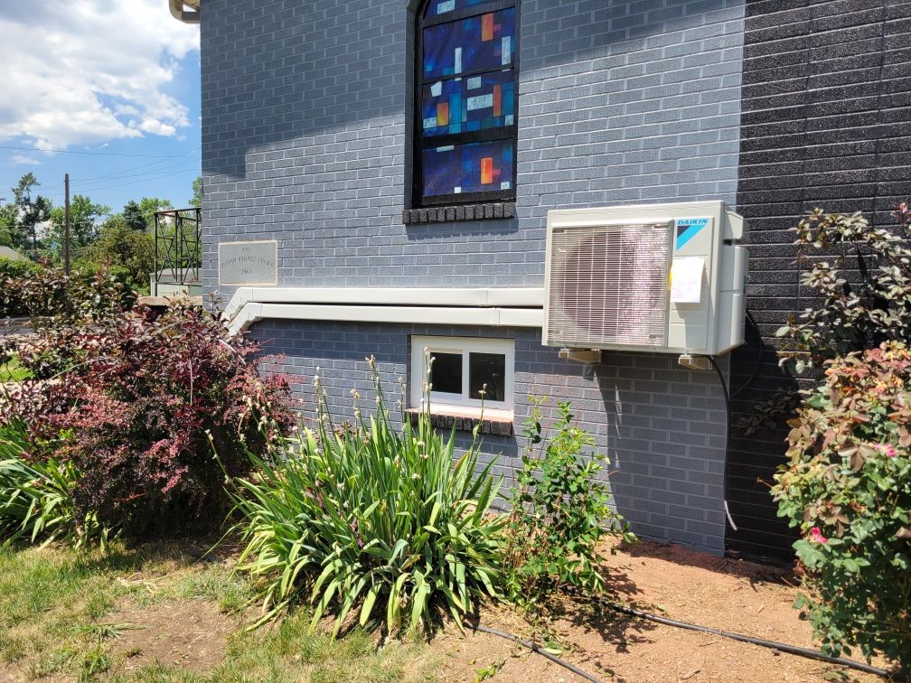 Exterior wall with air conditioner, window, and colorful stained glass, surrounded by bushes and grass.