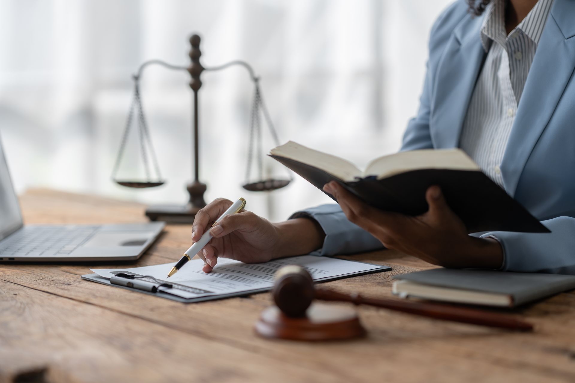 Person in a blue blazer reviewing documents and a book at a desk with a gavel and scales of justice.