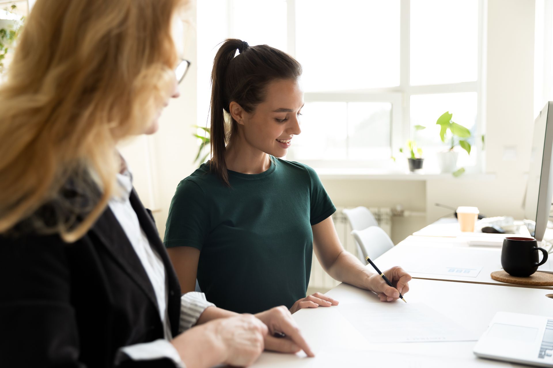 Two women at a desk, one points, the other signs document. Bright office, white walls, natural light.