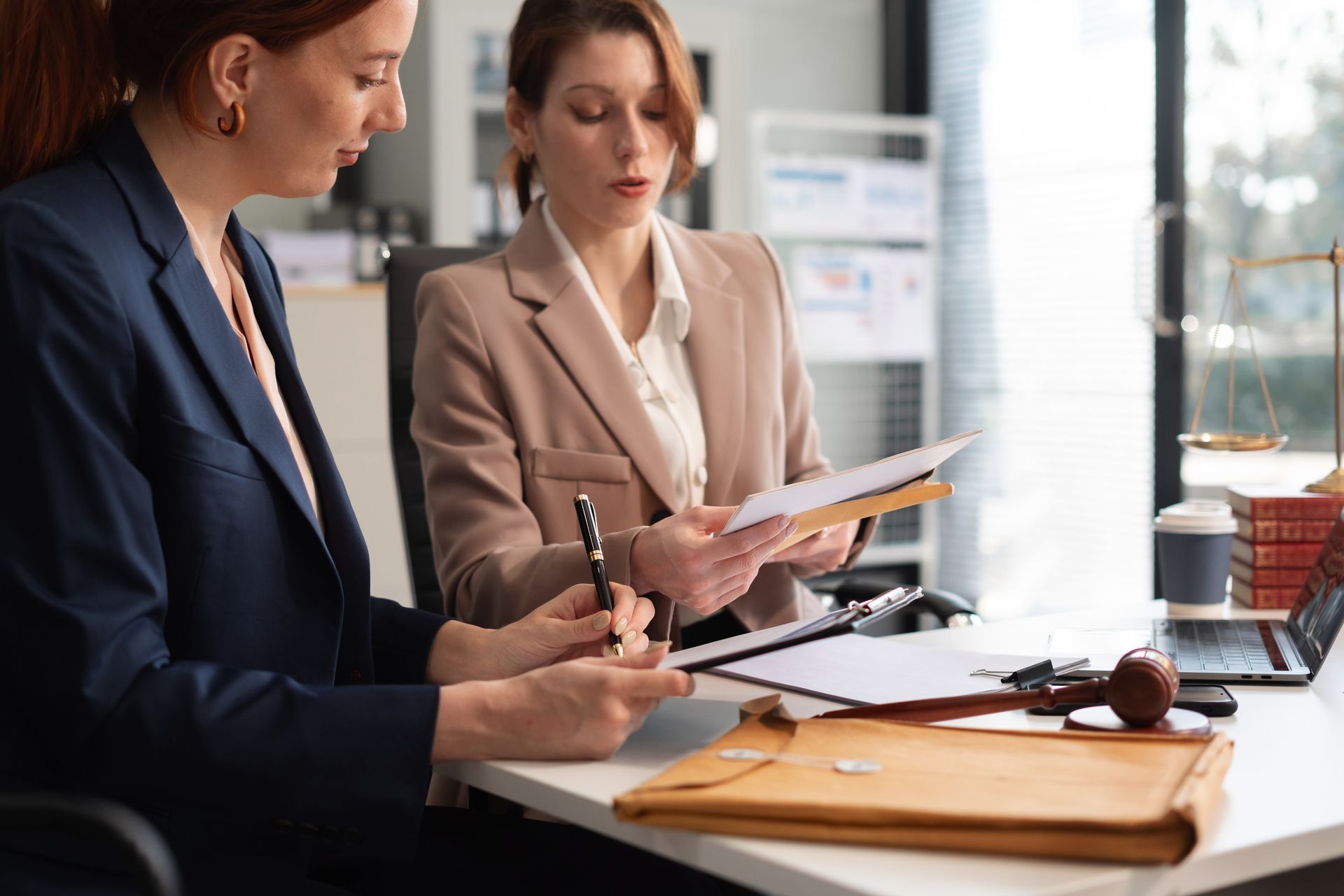 Two women reviewing documents at a desk in an office setting. One writes, the other holds papers.