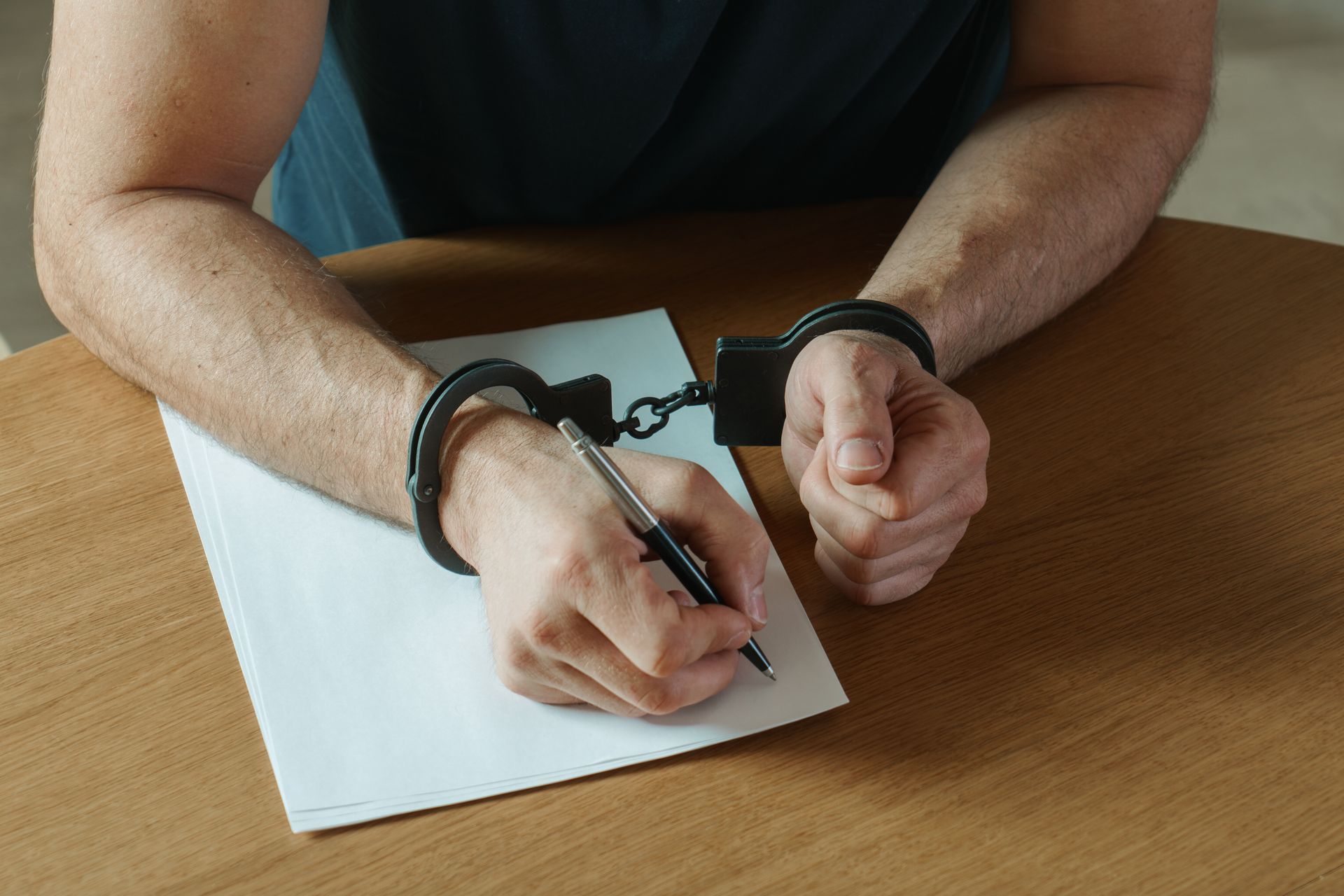 Hands in handcuffs writing on paper at a wooden table.