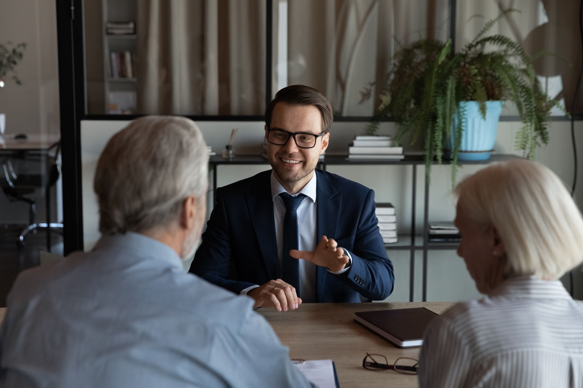 A man in a suit gestures, speaking to an older couple across a table in an office.