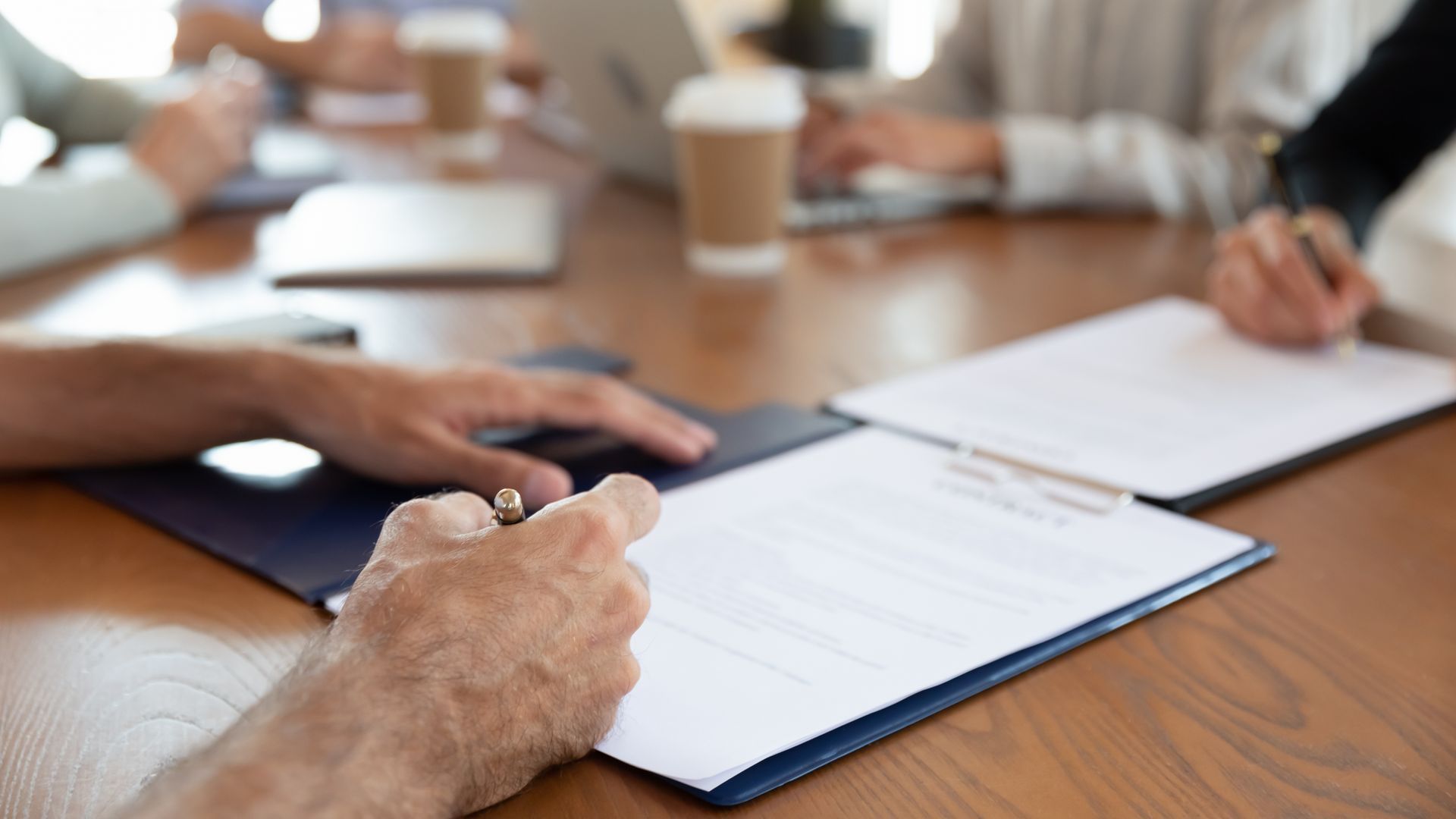 Hands signing documents around a wooden conference table. Other people work on computers.