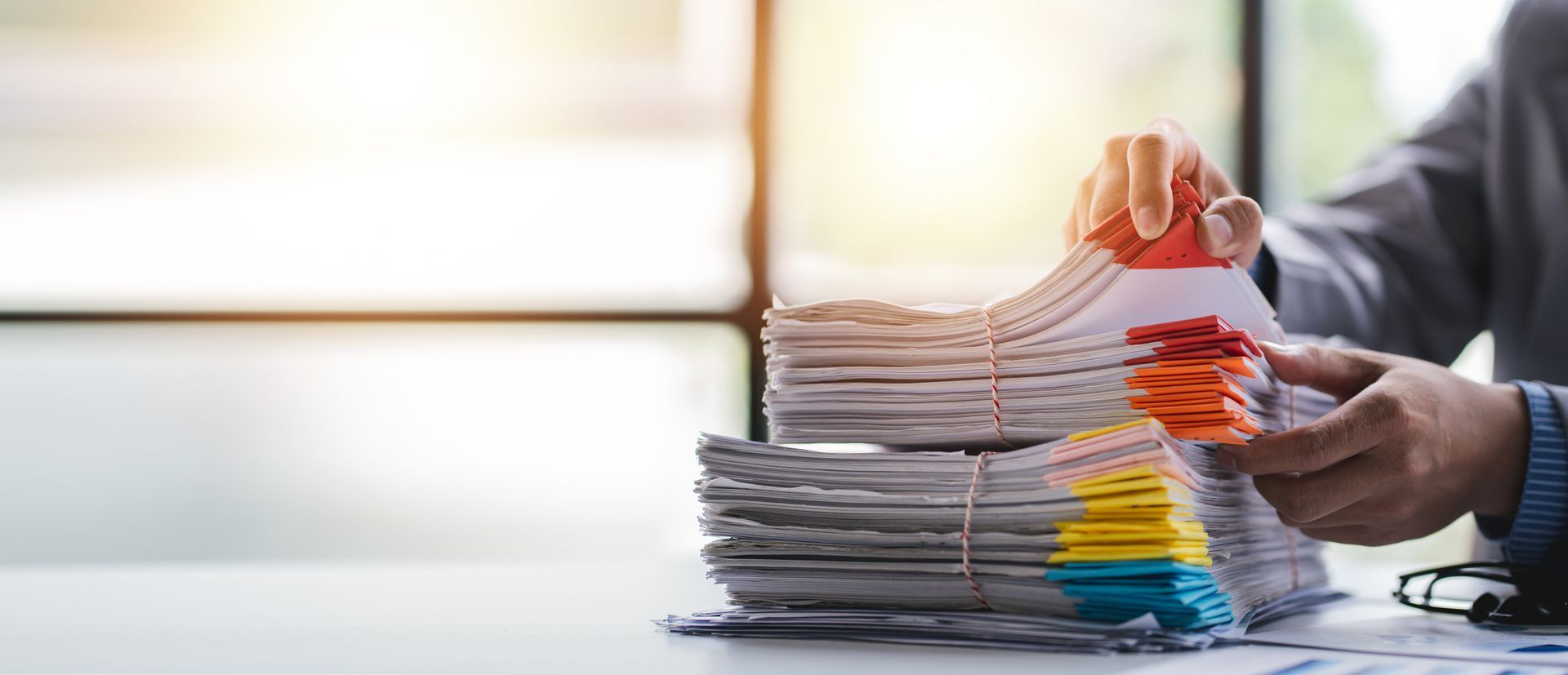 Person sorts through a large stack of papers, some color-coded, on a white desk near a window.