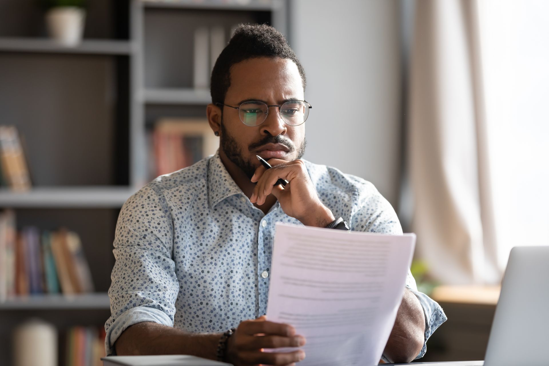 Man in glasses and patterned shirt looks at document, holding pen to chin, with a laptop and bookshelf in the background.