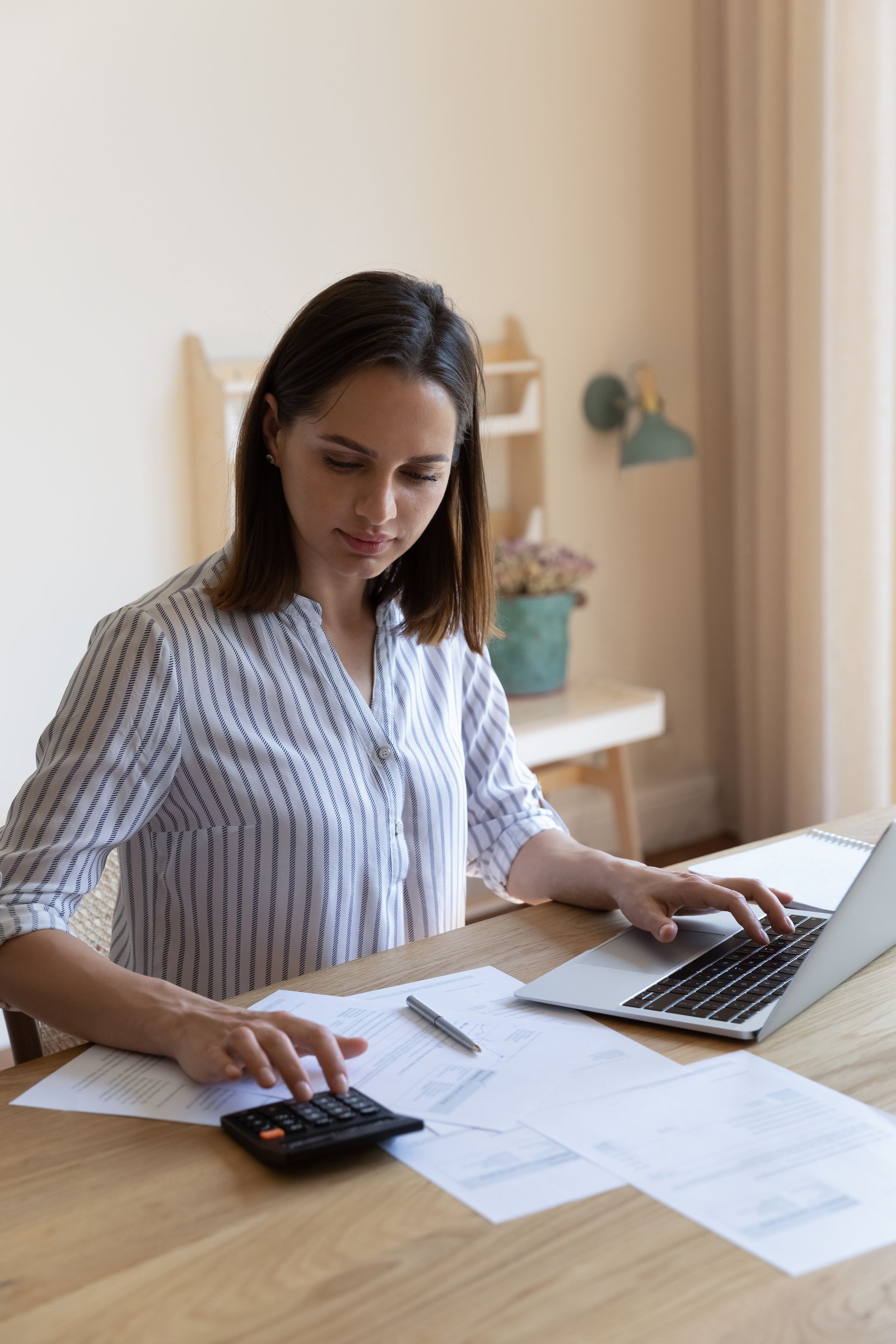Woman working on laptop, using a calculator and looking at papers on a wooden table.