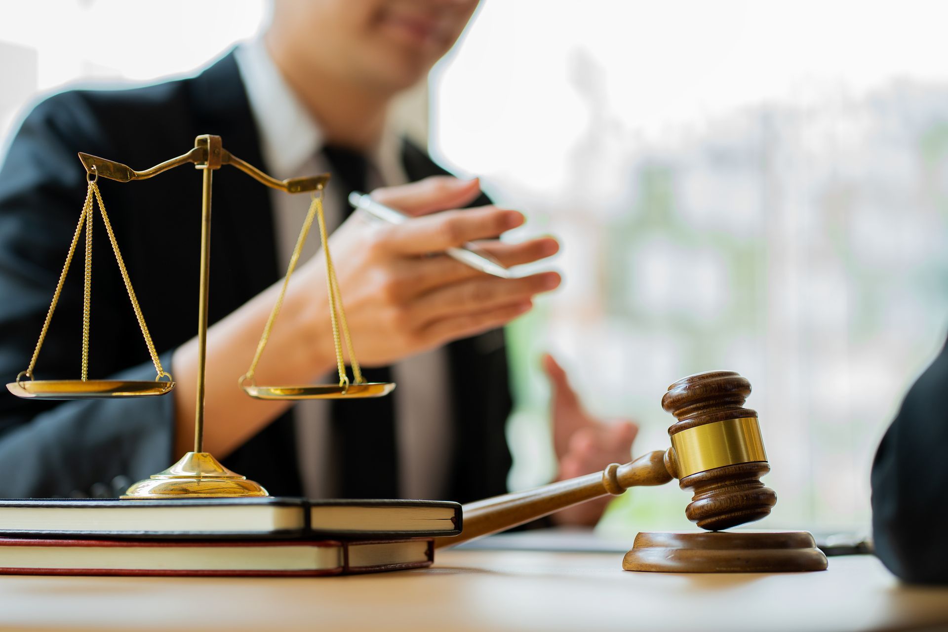 Scales of justice and gavel on a desk, a person in a suit gesturing with a pen in the background.