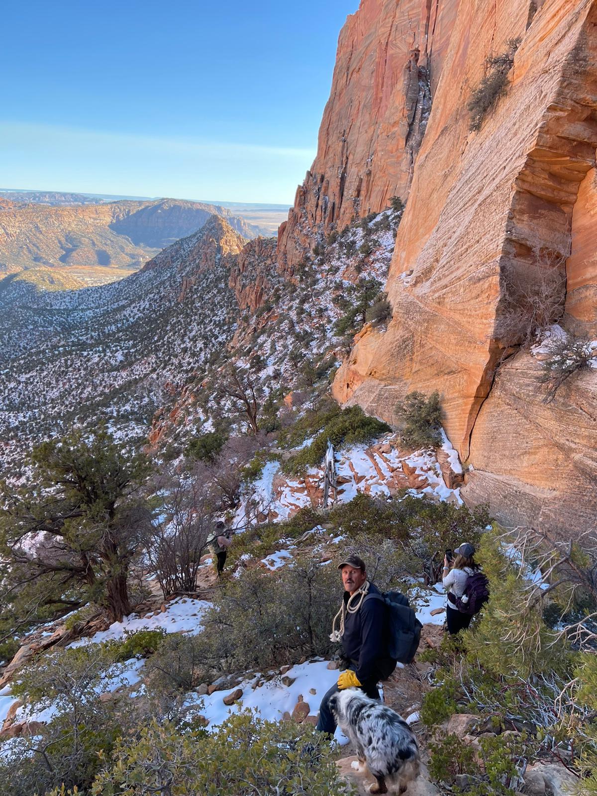 Hikers and their dog navigating a snowy trail along a cliffside with expansive views near Zion Glamping Adventures.