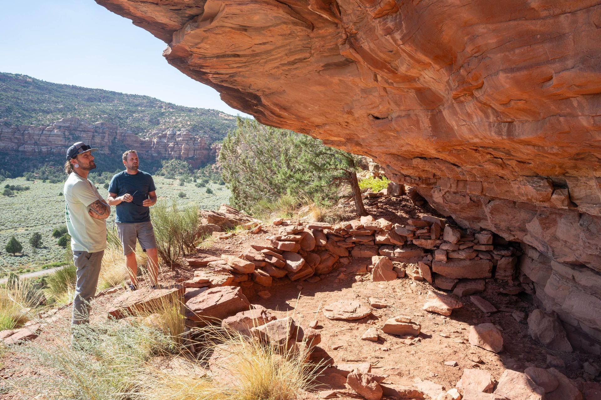 Two men exploring ancient ruins under a red rock overhang, with sweeping views of the surrounding landscape.