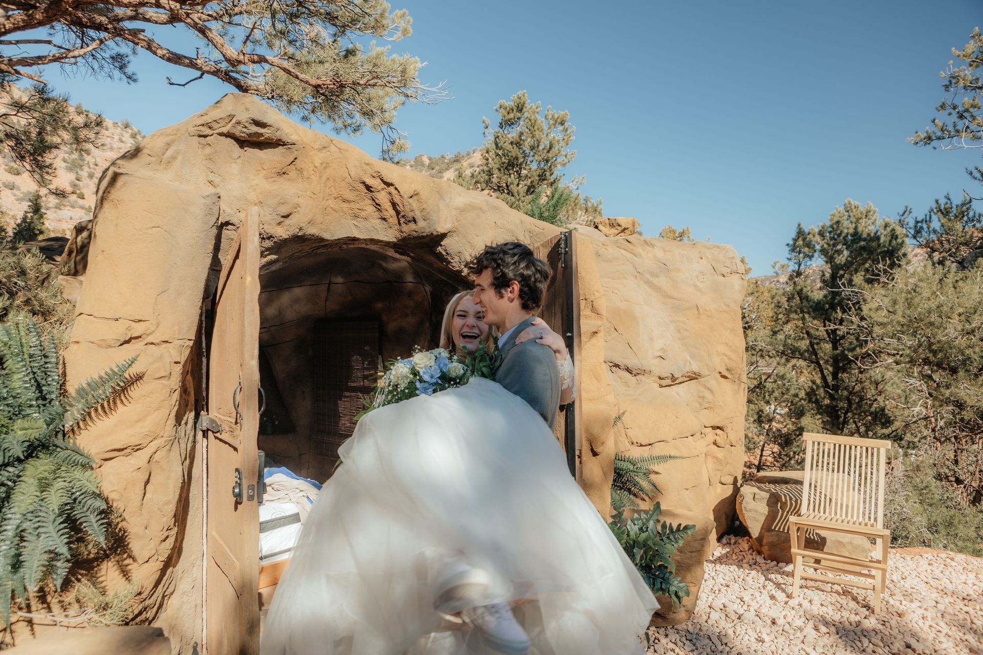 Newlyweds laughing and celebrating in front of a rustic rock cave accommodation at Zion Glamping Adventures