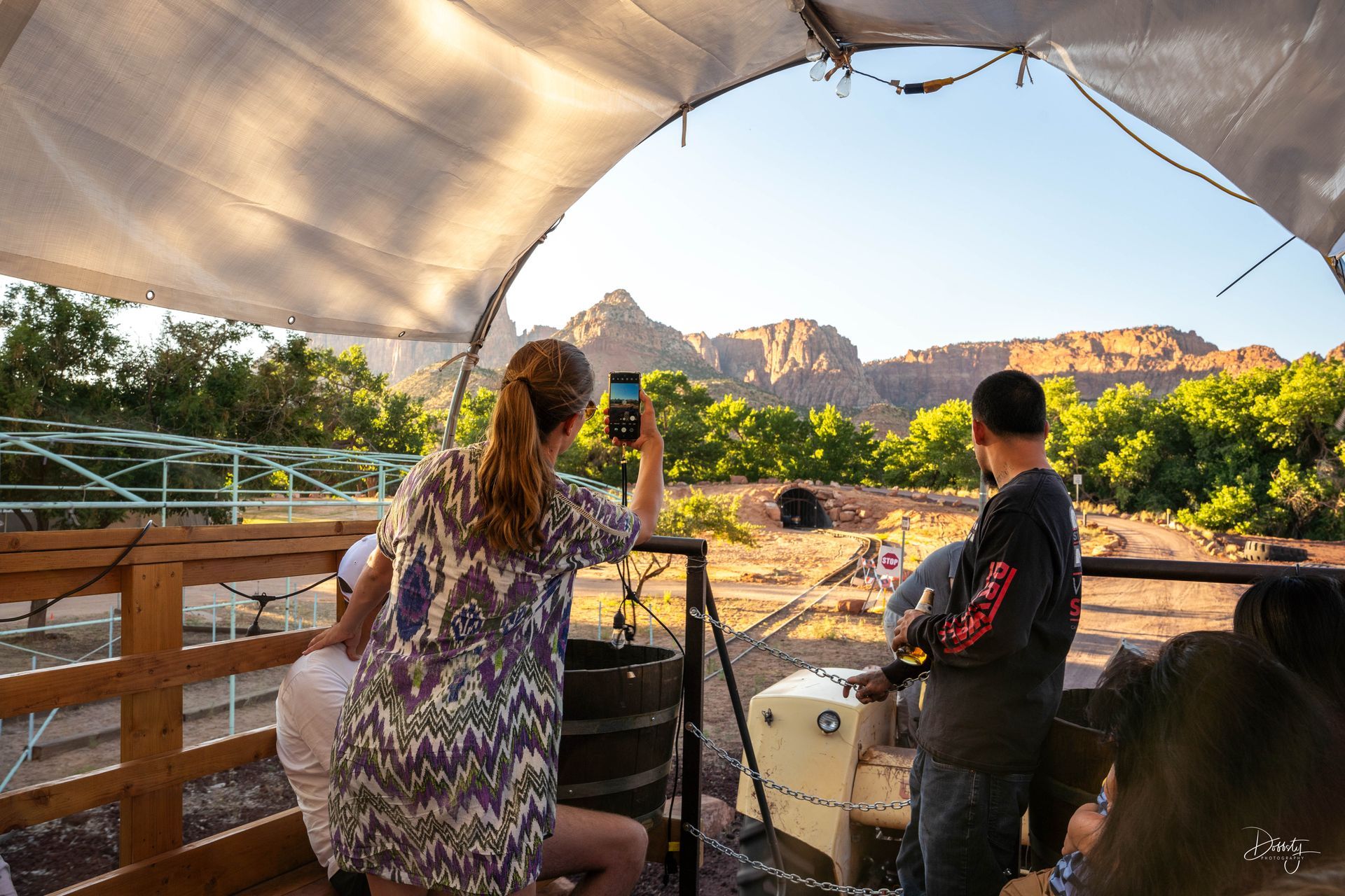 Tourists enjoying the view from a covered wagon, with a woman taking a photo of the mountainous landscape.