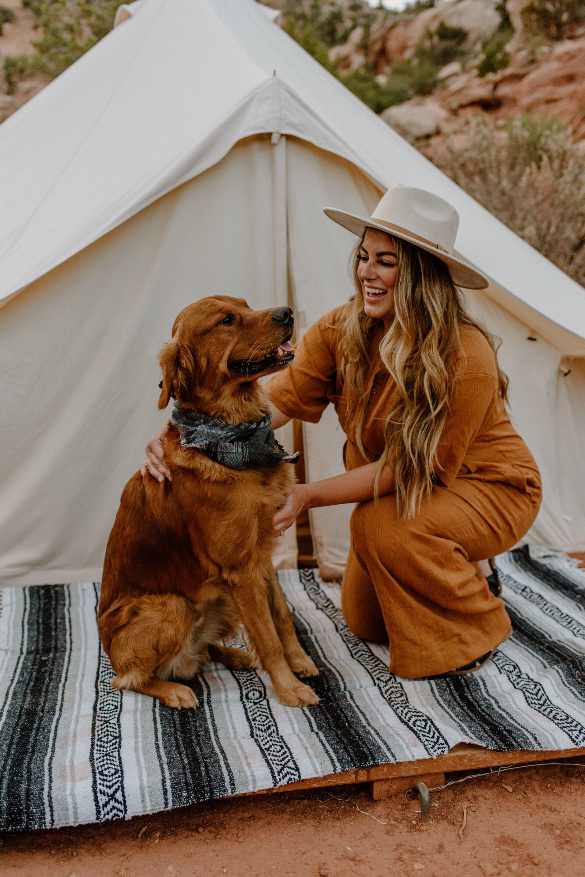 A woman in a hat smiles while petting a golden retriever in front of a glamping tent.
