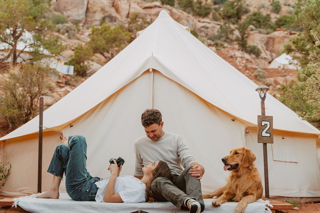 A couple and their golden retriever relax on a blanket outside their glamping tent, enjoying the outdoors.