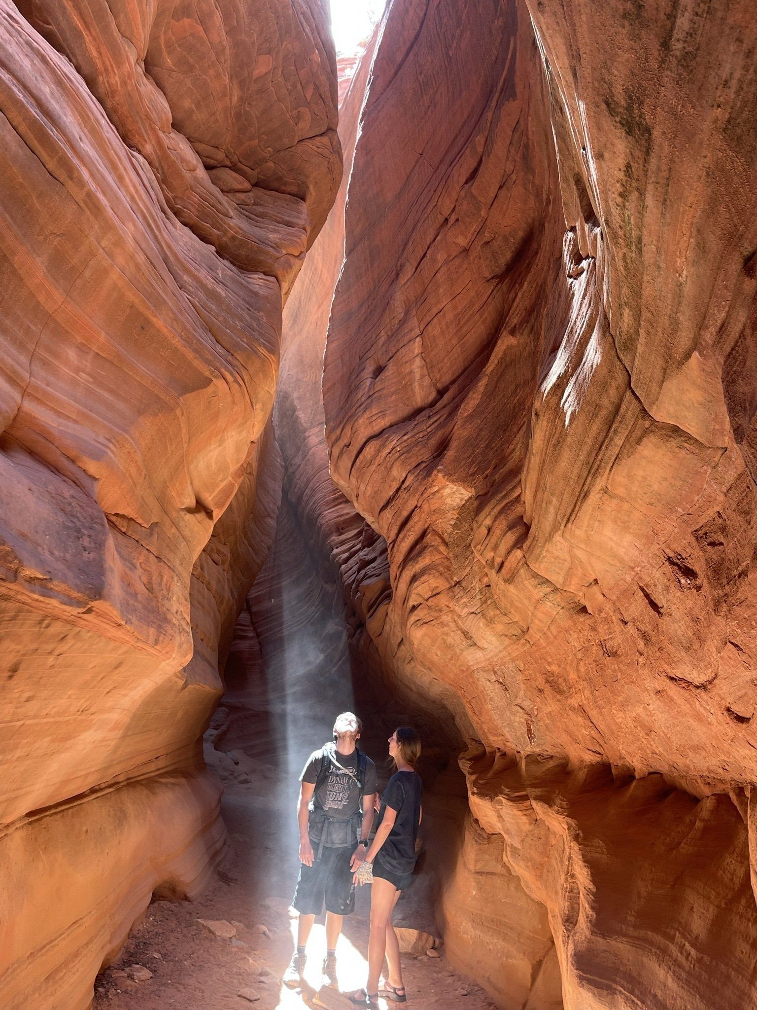 Couple standing inside a narrow slot canyon, illuminated by a beam of sunlight filtering through the red rock walls.